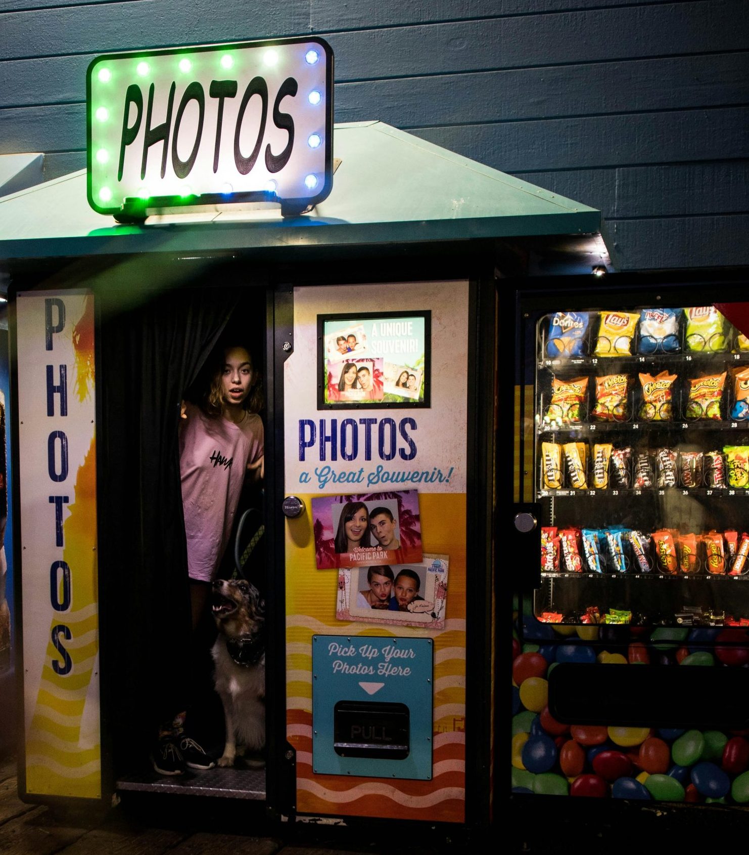 A woman and a dog standing inside a vintage photobooth at night surrounded by vending machines.