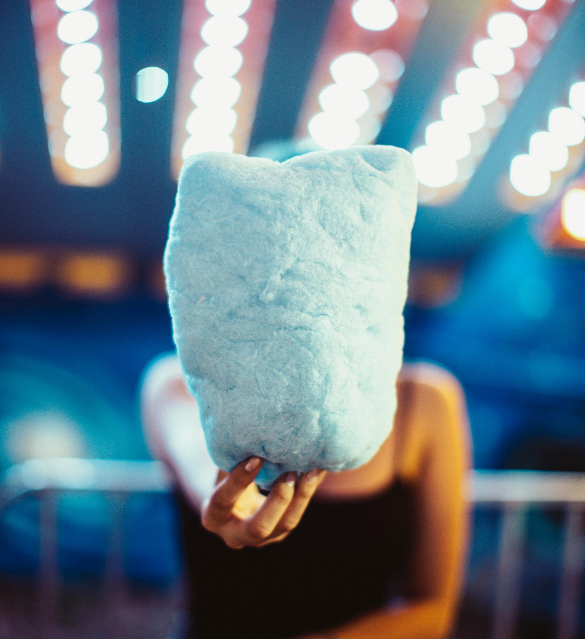 A person holds blue cotton candy with vibrant lights in the background at a night event.