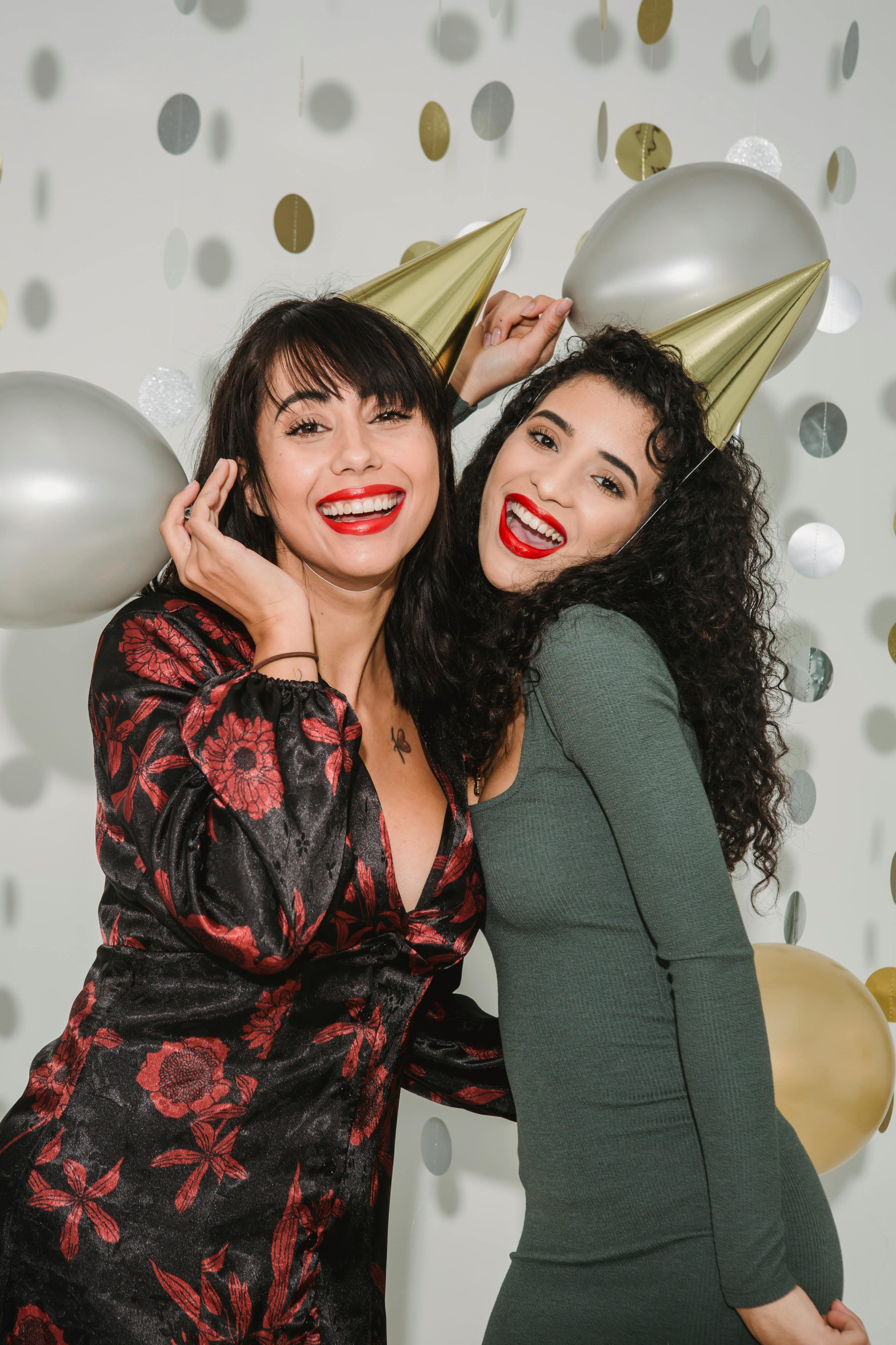 Positive ethnic woman embracing daughter in cone cap while celebrating holiday together in room with shiny decorations and balloons